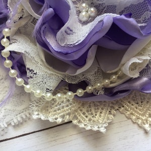 May include: A purple and white flower made of fabric with a pearl and lace trim.