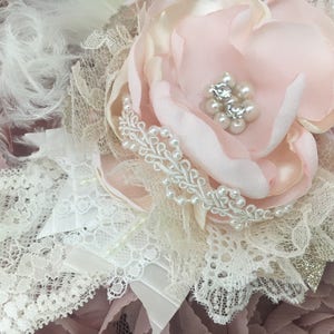 以下が含まれることがあります： 繊細な花のヘッドピースのクローズアップ。ヘッドピースは、パールとラインストーンの中心部を持つ大きな薄いピンク色の布の花、白いレース、羽、リボンに囲まれています。パールビーズのアクセントが詳細を追加します。