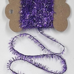 May include: A spool of purple tinsel yarn wrapped around a brown cardboard bobbin. The yarn has a metallic, reflective texture with fine, bristly fibers extending from a central core, and a loose length of the same purple tinsel yarn extends below.