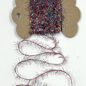 May include: A spool of colorful tinsel yarn, featuring a mix of pink, purple, gold, and teal strands. The yarn is wound around a brown cardboard spool, with some yarn unwound and lying below. Ideal for crafting and holiday decorations.