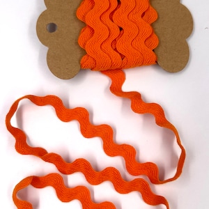 May include: Orange ric rac ribbon on a brown cardboard spool. The ribbon is wavy and has a textured appearance. The spool is on a white background. This ribbon is suitable for sewing and craft projects.