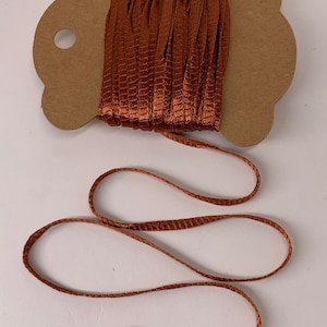 May include: A spool of copper-colored ribbon with a woven texture. The ribbon is unwound, with a loop of the ribbon extending from the spool. The spool is attached to a brown cardboard backing.