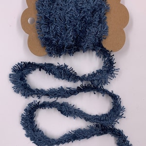 May include: A spool of navy blue fringe trim. The trim is fluffy and textured, with a wavy, fringed edge. The spool is attached to a brown cardboard backing with a scalloped edge. The trim is unwound and lies in a wavy line.