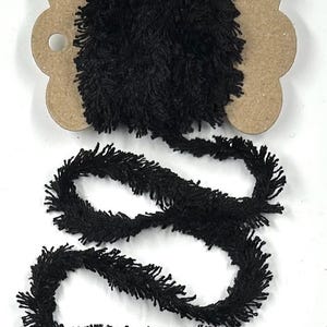 May include: A length of black fringe trim wrapped around a scalloped-edge brown cardboard spool. The trim features a dense, fuzzy texture with short, protruding fibers along the entire length of the cord, creating a consistent appearance.