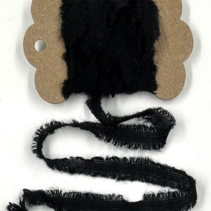 May include: A spool of black frayed ribbon. The ribbon is tightly wound on a cardboard spool with a decorative edge. The ribbon has a textured, fringed edge and is unspooled in a length on a white background.