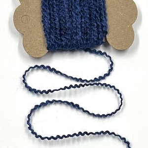 May include: A spool of navy blue decorative trim with a wavy, textured design. The trim is wound around a brown cardboard spool with a unique, scalloped edge. The trim is unwound, creating a flowing, curved line.