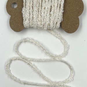 May include: A spool of off-white, textured yarn. The yarn is wound around a brown cardboard spool with a decorative shape. A length of the yarn extends from the spool, creating a looping pattern. The yarn appears soft and slightly fuzzy.