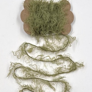 May include: A spool of olive green fringe trim sits atop a white surface. The trim is made of a soft, textured material and is partially unwound, revealing strands of the same color. The spool is made of a light brown cardboard.