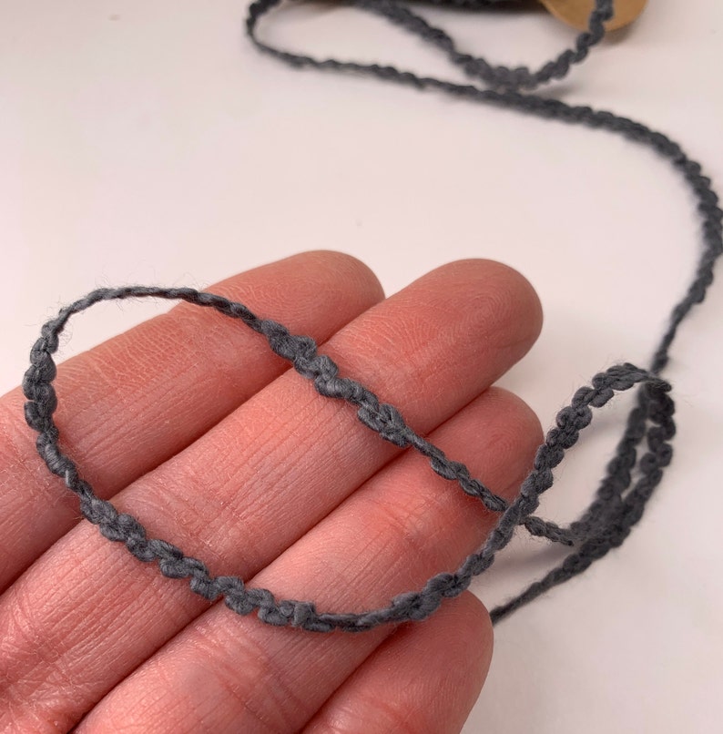 Puede incluir: Una cinta decorativa gris con borde festoneado. La cinta est&aacute; hecha de un material suave y tejido. La cinta se muestra sobre un fondo neutro. Adecuada para diversos proyectos de artesan&iacute;a y costura.