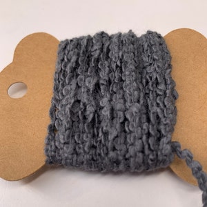 Puede incluir: Una bobina de hilo gris oscuro texturizado. El hilo est&aacute; enrollado firmemente en una bobina de cart&oacute;n marr&oacute;n con un borde decorativo. El hilo tiene una textura en bucle y nudosa, y un hilo se extiende desde la bobina.