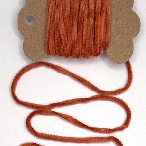 May include: A spool of orange-brown velvet yarn. The yarn is wound around a cardboard spool with a decorative, three-lobed design on each end. The yarn is soft and fluffy, with a slight sheen.