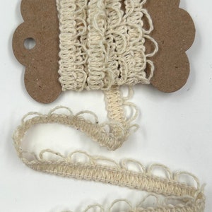 May include: Off-white, open-weave trim on a cardboard spool. The trim has a scalloped edge and is unraveled in a length below the spool. The trim is made of a soft, natural fiber, likely cotton or linen.