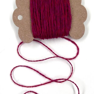 May include: A spool of vibrant magenta-colored twine. The twine is wound around a cardboard spool with a decorative edge. A length of the twine extends from the spool, creating a looping pattern against a white background. Ideal for crafting and decorative projects.