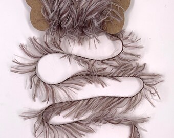 May include: A spool of decorative feather trim in shades of gray and white, attached to a brown cardboard backing. The trim is arranged in a wavy pattern, showcasing its texture and color variations. Ideal for craft projects.