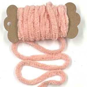 May include: A spool of light pink chenille yarn wrapped around a brown cardboard holder with scalloped edges. The soft, fuzzy yarn is wound tightly around the center, with a loose strand extending downward in a series of loops.