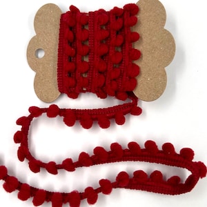 May include: A spool of red pom-pom trim. The trim is a deep red color and features small, round pom-poms along the edge. The trim is wound around a cardboard spool with a scalloped edge. The trim is unwound and lying on a white surface.