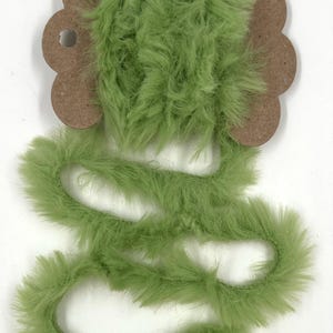 May include: A spool of light green faux fur trim. The trim is a soft, fluffy material with a slightly wavy texture. The spool is attached to a brown cardboard backing with a flower-like shape.