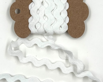 May include: White wavy ribbon on a brown cardboard spool. The ribbon has a textured, ruffled edge and is unspooled at the bottom. The spool has a decorative shape with a hole for hanging. Ideal for crafts and sewing projects.