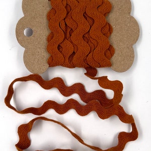 May include: A spool of brown wavy ribbon. The ribbon is a rich, warm brown color and has a textured, ruffled edge. The ribbon is wound around a cardboard spool with a decorative shape. The ribbon is unwound in a wavy pattern.