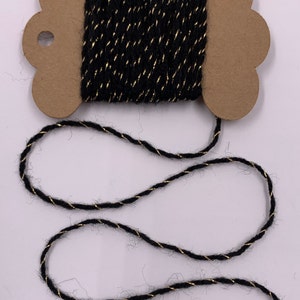 May include: A spool of black twine with a gold metallic thread woven throughout. The twine is wound around a brown cardboard spool with a decorative edge. The twine is unwound and lies on a white surface.