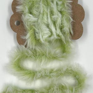 May include: A length of fluffy, light green and white faux fur trim, attached to a brown cardboard tag with a hole for hanging. The fur is soft and textured, ideal for craft projects or costume making.