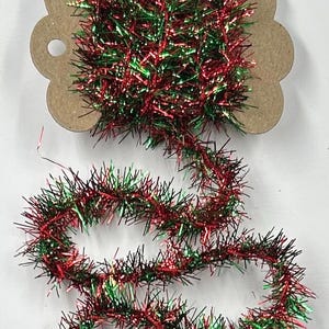 May include: A spool of metallic tinsel trim featuring a vibrant mix of red and green strands. The tinsel is wrapped around a scalloped-edge brown cardboard bobbin, with a portion of the shiny, reflective garland extending downward in loose coils.