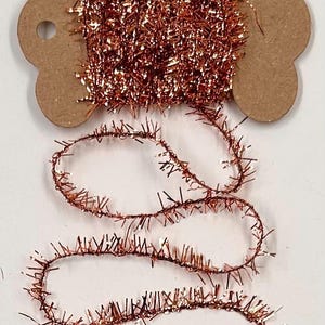 May include: Copper-colored tinsel ribbon wound on a brown cardboard spool. The tinsel has a metallic sheen and is arranged in a decorative pattern. The ribbon is unwound in a looping design, suitable for crafts and decorations.