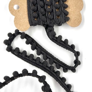May include: A spool of black pom-pom trim with a decorative edge. The trim is made of a soft, textured material and is displayed on a brown cardboard spool. The trim is unraveled, showing the pom-pom detail.