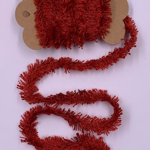 May include: A spool of rust-colored fringed trim. The trim is made of a soft, textured material and is wound around a cardboard spool. The trim is unwound, creating a flowing, decorative element. Ideal for craft projects.