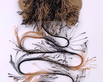 May include: A spool of decorative yarn in shades of black, brown, and tan. The yarn has a textured, fringed appearance, with strands extending from the main body. The yarn is wound around a brown cardboard spool, suitable for crafts and textile projects.