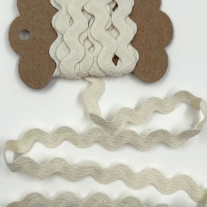 Puede incluir: Cinta ondulada de color crema sobre una bobina de cartón marrón. La cinta está desenrollada, mostrando su patrón de zigzag texturizado. La cinta está hecha de un material suave y tejido, ideal para manualidades y costura.