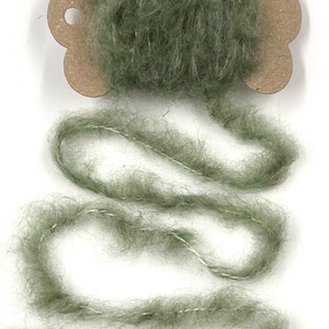 May include: A spool of sage green yarn with a fluffy texture and a thin white thread running through it. The yarn is displayed against a white background, with a brown cardboard tag attached to the spool. Ideal for knitting and crochet projects.