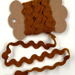 May include: A spool of brown wavy ribbon. The ribbon is a warm, earthy brown color and has a textured, wavy design. The ribbon is wound around a brown cardboard spool with a decorative edge. The ribbon is unwound from the spool.