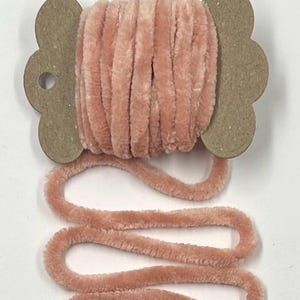 May include: A spool of soft, peach-colored velvet yarn. The yarn is wound around a cardboard spool with a decorative, scalloped edge. The yarn is unwound in a wavy pattern, showing its plush texture.