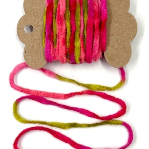 May include: A spool of colorful yarn in shades of pink, fuchsia, and green. The yarn is fluffy and textured, with a soft, velvety appearance. The spool is made of cardboard.