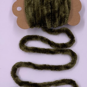 May include: A spool of olive green velvet yarn. The yarn is thick and plush, with a soft, textured appearance. The yarn is unwound, revealing a length of the same material. The spool is attached to a brown cardboard backing.