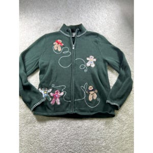 May include: A dark green zip-up jacket featuring long sleeves and a high collar. The front is decorated with embroidered white swirling lines and five appliquéd gingerbread figures in various plaid and solid fabric patterns with red accents.