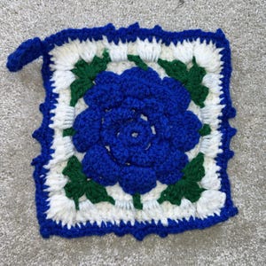 May include: A square, handmade crochet potholder featuring a large blue rose in the center. The rose is surrounded by white and green accents, with a blue scalloped border and a small loop for hanging. The potholder is made of yarn.