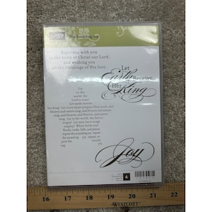 May include: A clear plastic stamp storage case labeled The Sounding Joy by Stampin' Up. The insert features black text with lyrics to Joy to the World and the phrases Let Earth Receive Her King and Joy. A wooden ruler is visible at the bottom.
