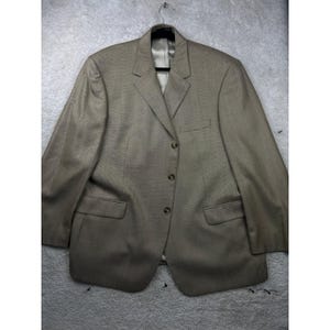 May include: A tan blazer featuring a three-button front closure, notched lapels, and two flap pockets at the waist. The garment includes a single chest pocket and long sleeves, presented on a black hanger against a neutral background.