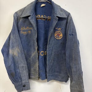 Peut inclure: Une veste en velours côtelé bleu avec une fermeture éclair frontale, un col pointu et des manches longues à poignets boutonnés. La poitrine porte le nom brodé Shelton Leggett et un écusson circulaire FFA Vocational Agriculture.