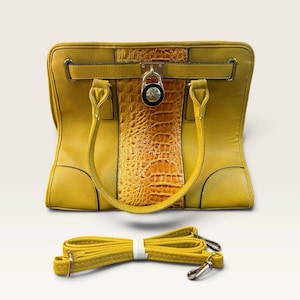 May include: A mustard yellow handbag featuring a central vertical panel with a textured crocodile skin pattern. The bag includes two top handles, a horizontal strap with a silver-toned metal padlock closure, and a detachable matching shoulder strap.