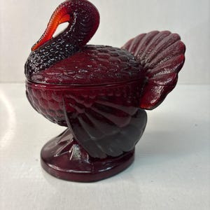 May include: A deep red, translucent glass turkey-shaped covered dish. The piece features intricate molded feather textures across the body, wings, and tail, with a curved neck and head forming the lid handle, resting on a circular base.