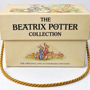Puede incluir: Una caja de almacenamiento rectangular de color crema con el texto The Beatrix Potter Collection e ilustraciones de conejos. La caja incluye un asa de cuerda trenzada dorada y el texto The Original and Authorized Editions impreso.