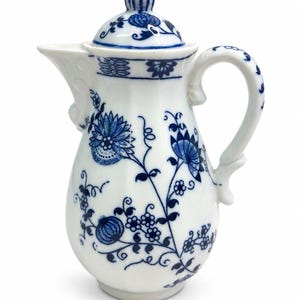 May include: A white porcelain coffee pot featuring a blue floral pattern with intricate botanical motifs, a curved handle, a pointed spout, and a matching lid with a finial. The surface is decorated with detailed blue painted flowers.