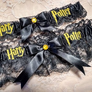 Może przedstawiać: Dwie czarne koronki do pończoch z czarnymi satynowymi kokardkami i złotymi cekinami. Koronki są wyszyte tekstem "Harry Potter" w kolorze żółtym.