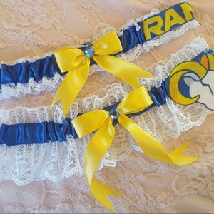 Könnte beinhalten: Zwei blaue und weiße Strumpfhalter mit gelben Schleifen und blauen Strasssteinen. Ein Strumpfhalter hat ein Rams-Logo und der andere sagt "Rams".