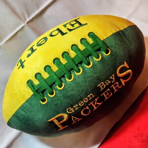 May include: A plush football-shaped pillow featuring a yellow and dark green color scheme. The surface displays embroidered text reading "Ebert" and "Green Bay PACKERS" in yellow thread. A central green lace-up detail mimics a football design.