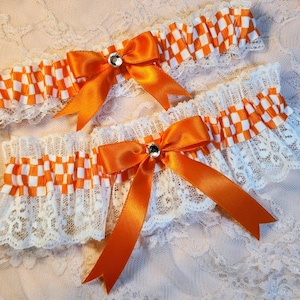 May include: Two elastic leg garters featuring a white and orange checkered ribbon trim layered over white floral lace. Each garter is adorned with a centered orange satin ribbon bow and a small, clear, round rhinestone accent at the center.