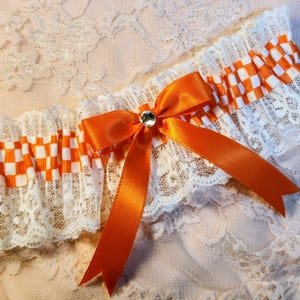 May include: A white lace garter featuring a ruffled trim with an orange and white checkered ribbon accent. A central orange satin bow is embellished with a single clear rhinestone at the knot, creating a decorative accessory with lace.